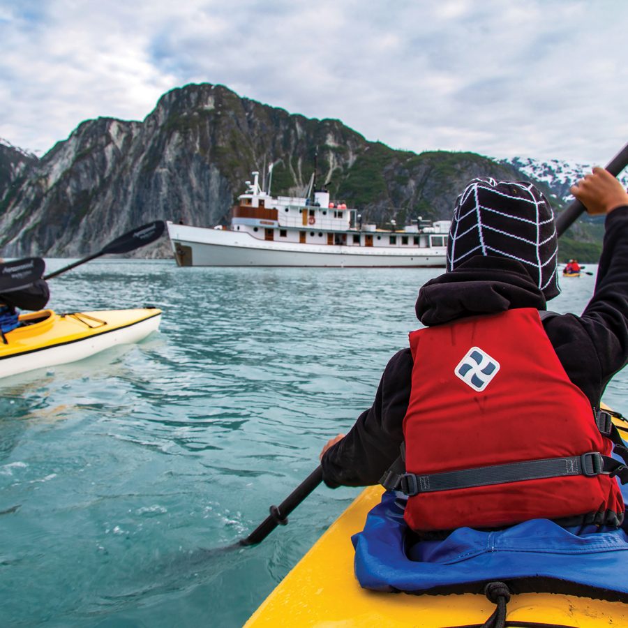 People paddle in kayaks near a small ship. Rocky mountains covered in snow surround the water. 