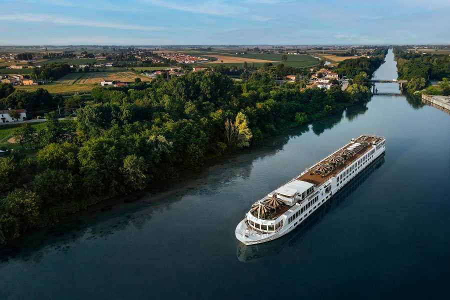 A long white river cruise ship travels along a quiet river surrounded by trees and fields.