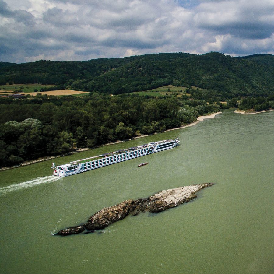 A long white river cruise ship travels along a green-tinted river surrounded by fields and woodland in Austria.