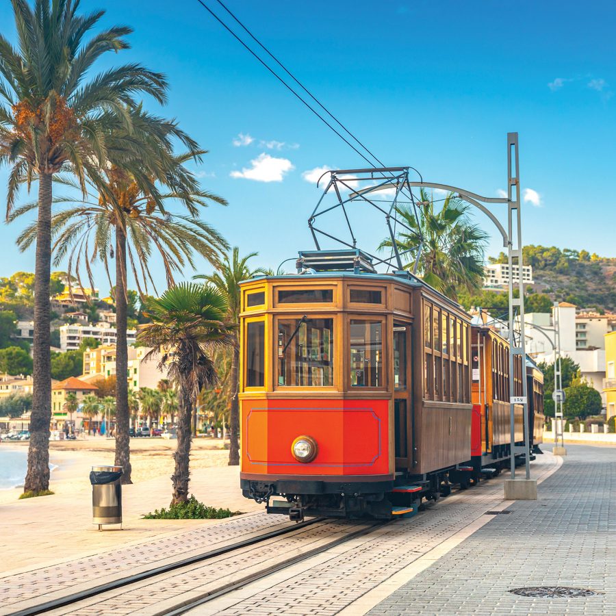 A old-fashioned red and gold tram passes a sandy beach lined with palm trees in Palma de Mallorca.