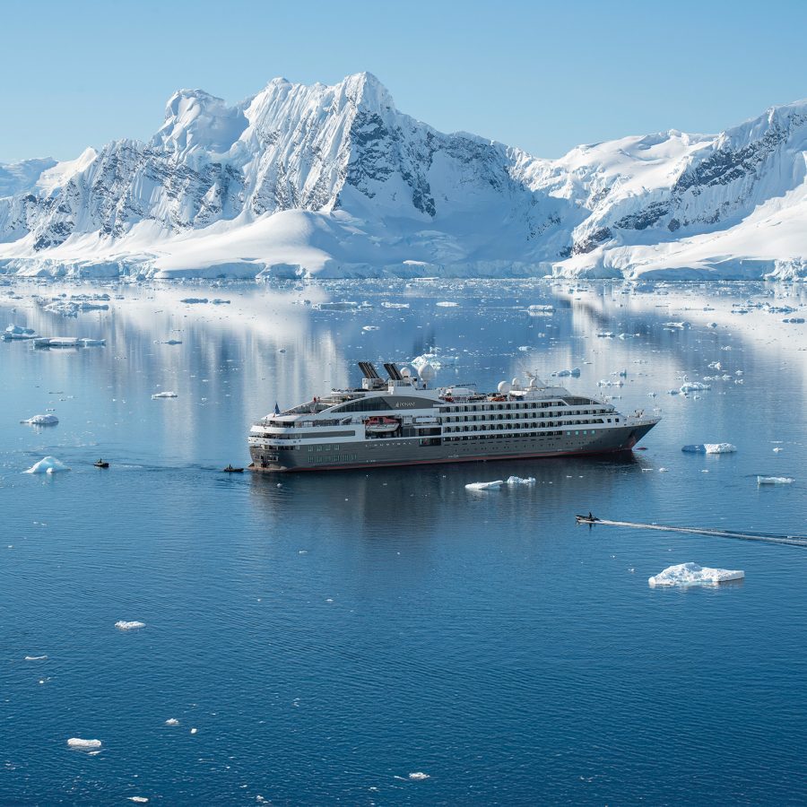 A large white cruise ship travels through water strewn with pieces of floating ice. In the background are rocky peaks covered in snow.