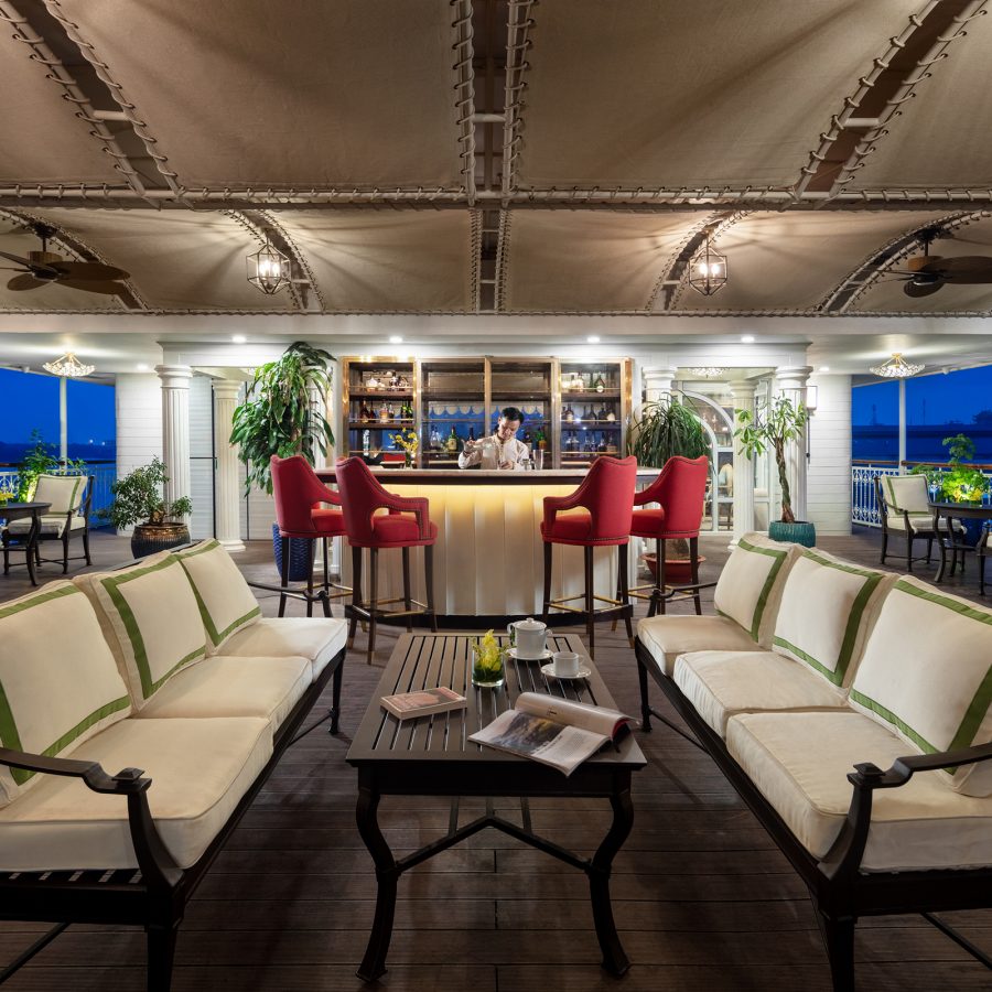 A bartender stands behind a spotlit bar on the open-plan deck of a cruise ship. 