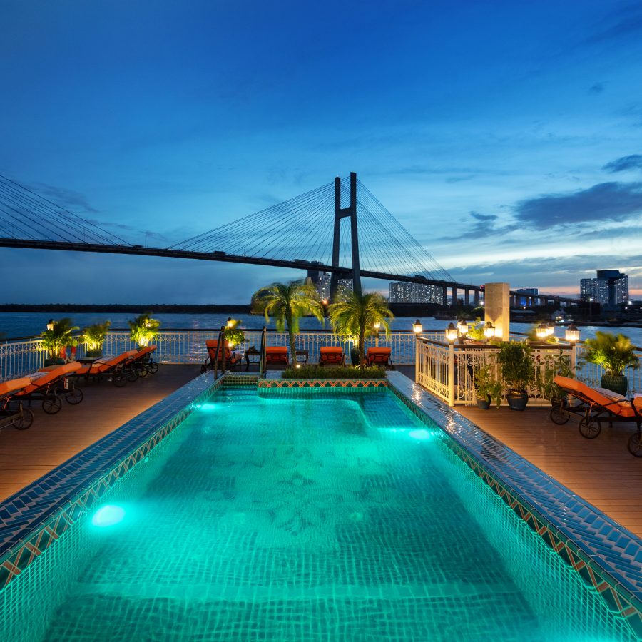 A swimming pool is illuminated by spotlights on the sundeck of a river cruise ship. In the background, the sun sets over a large suspension bridge.