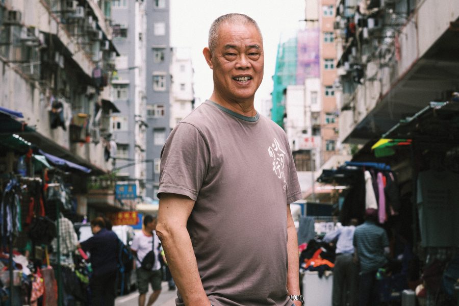 Ming Gor, or Chan Cheuk-ming, stands smiling on a street lined with stalls in Sham Shui Po.