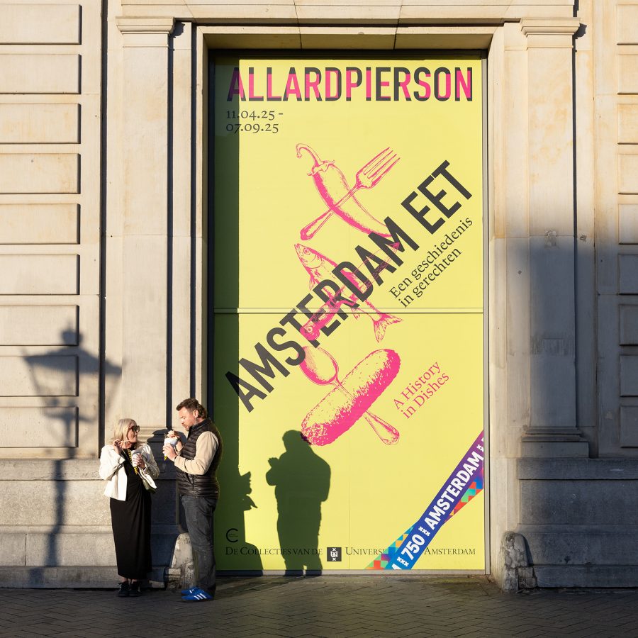 A streetview of two men standing infront of a poster of Amsterdam Eet on the a building wall with sunlight casting a shadow on the building in Amsterdam, Netherlands 