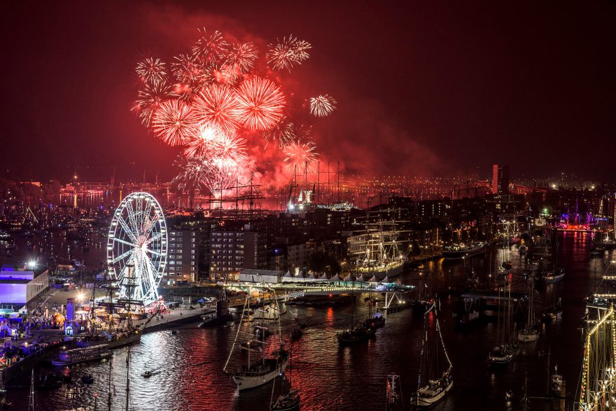 A view of tall historic ships and naval vessels with fireworks and a Ferris wheel in the background at IJ Harbour of Amsterdam, Netherlands at night