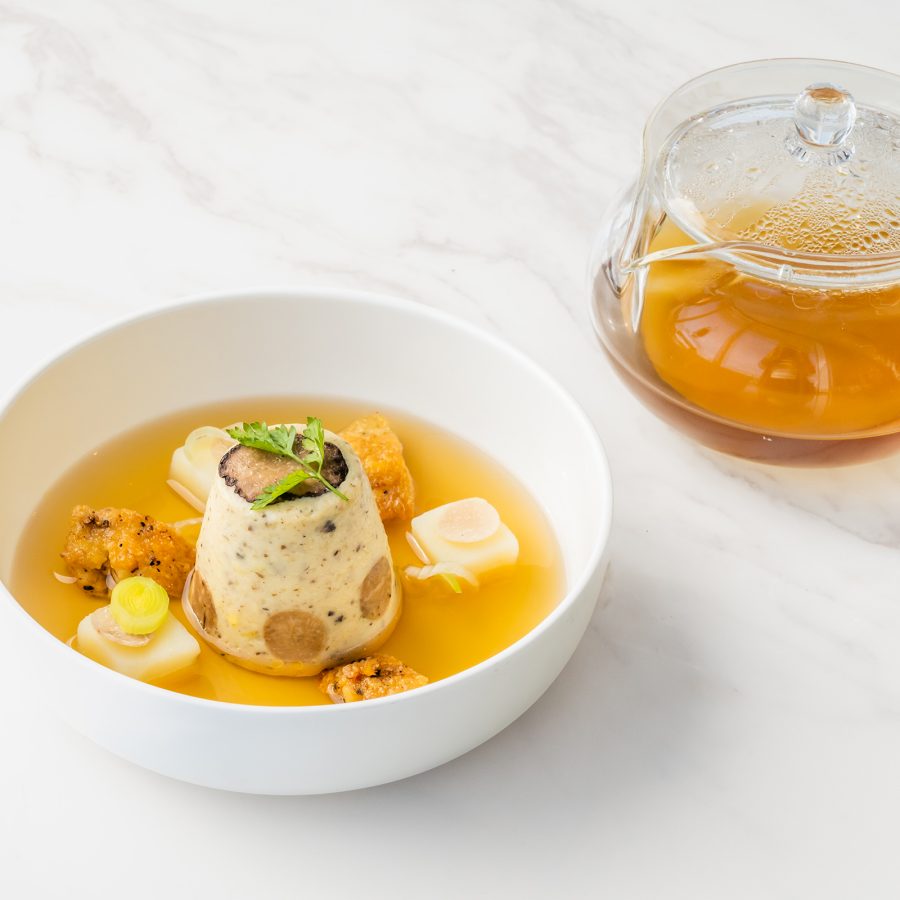 A white bowl of fois gras ravioli in duck consume and a side of broth in a glass pot served at Harbourside Grill