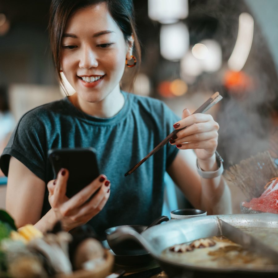A woman smiling at mobile phone, chopsticks in hand infront of hot pot 