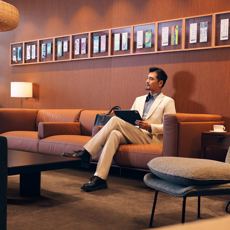 A man in a suit sitting on a sofa with a tablet at the Beijing Cathay Pacific lounge. William Lim’s boarding pass drawings are framed and hung on the wall above him.
