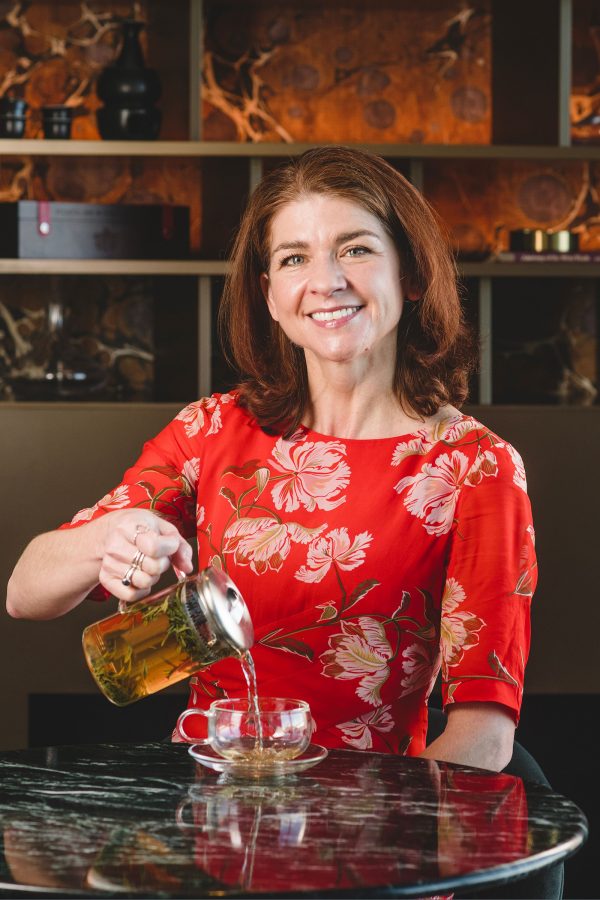 A woman in a red top pours a kettle of JING tea into a cup. 