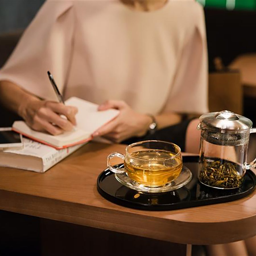 A woman writing in a notebook, with a cup of JING tea on the table. 