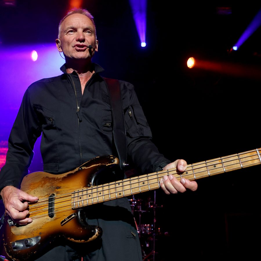 Sting performs on stage as stage lighting shines in multiple directions behind him.