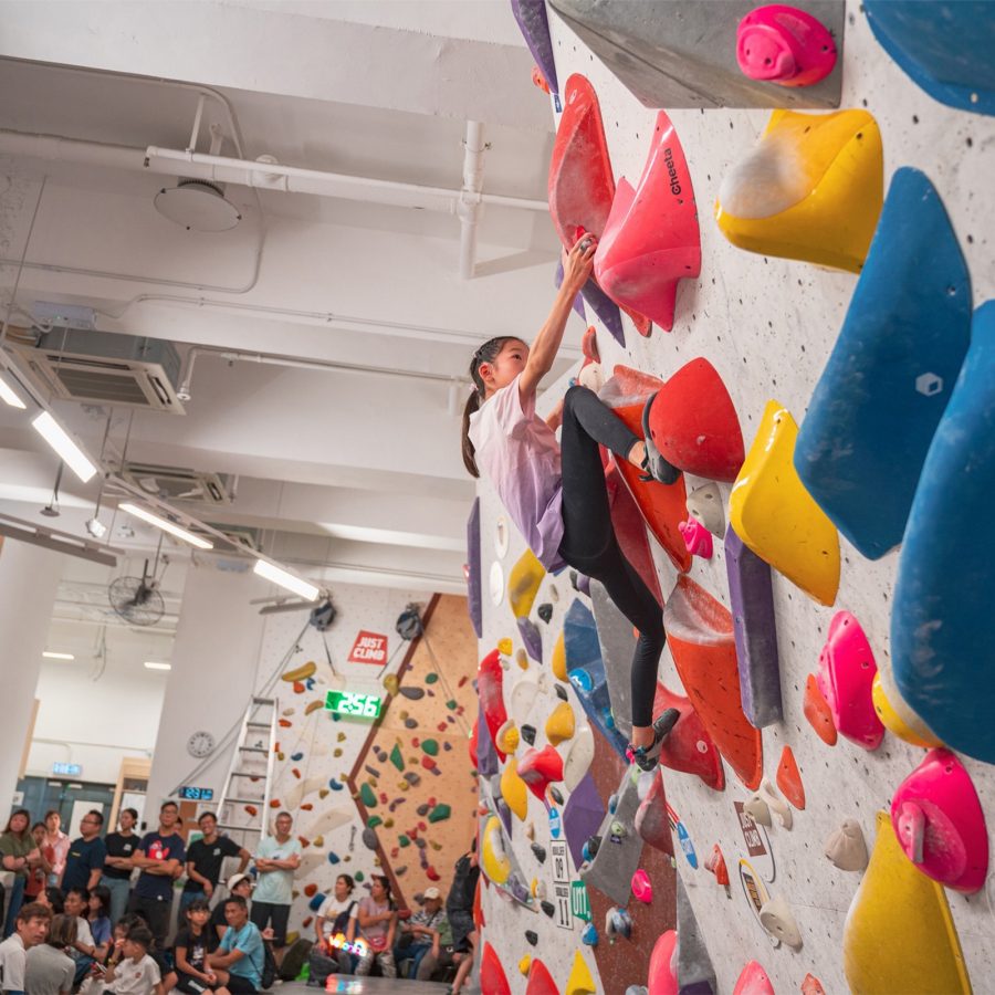 Interior view of a group of people watching a young woman bouldering at Just Climb. 