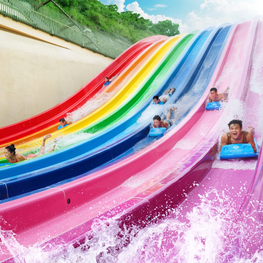 People lying face down on mats race down rainbow-coloured slides at Water World Ocean Park.