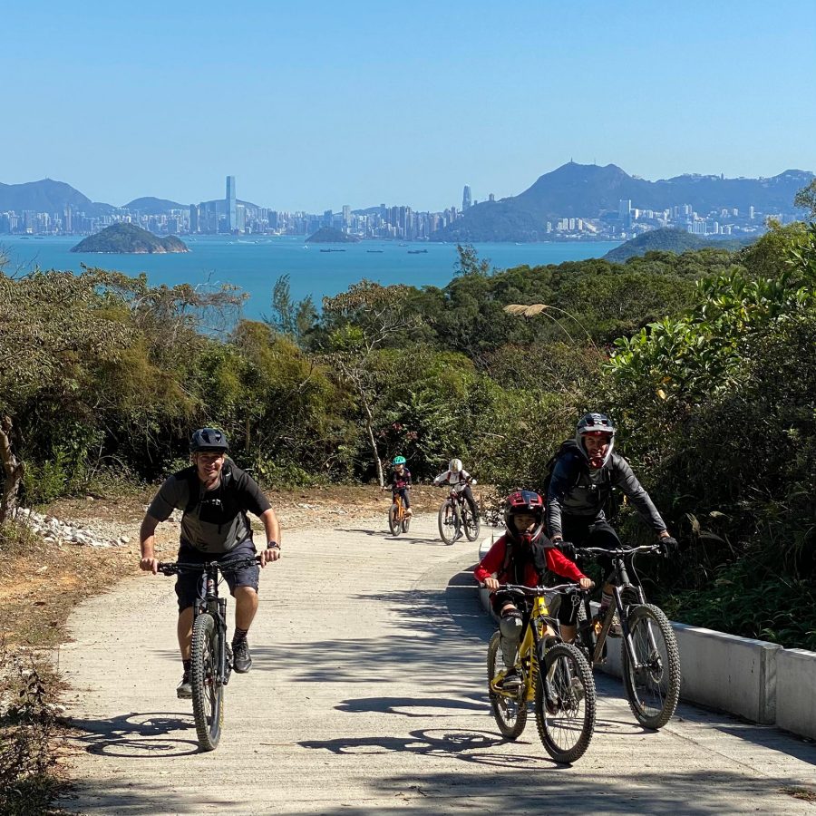 A group of adults and children riding bicycles on a path with a body of water and mountains in the background.