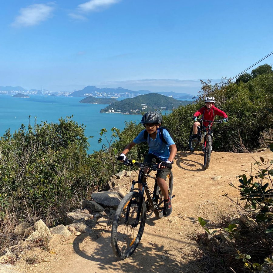 Two people riding bikes on a trail next to a body of water and mountains.