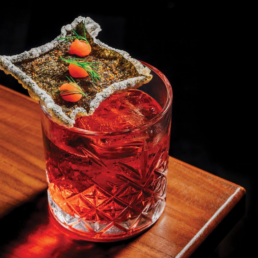 A Japanese-inspired red cocktail in a glass on a table, garnished with seaweed. 