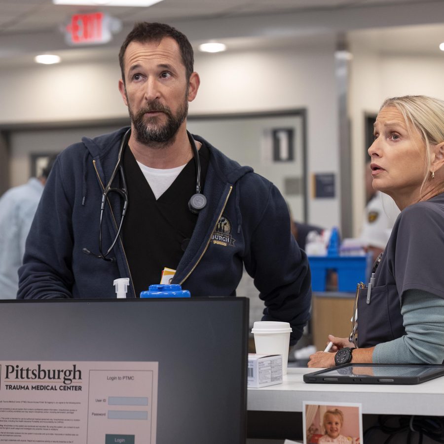 A man and woman dressed in medical scrubs stand next to a counter in a hospital, in a scene from The Pitt.