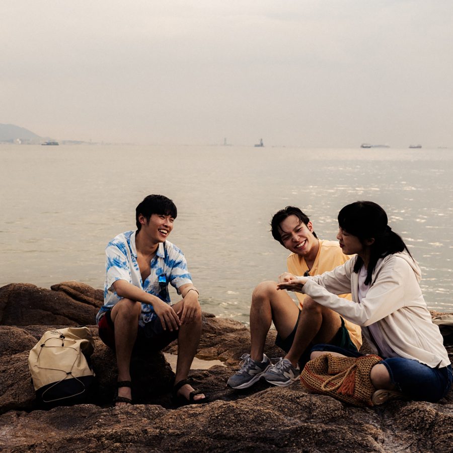 Three young people smile at each other while sitting on some rocks next to the sea in a scene from The Way We Talk.