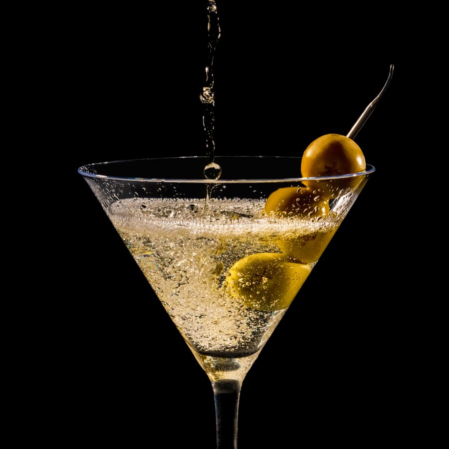 A martini being poured into a glass with olives on a stick against a black backdrop. 