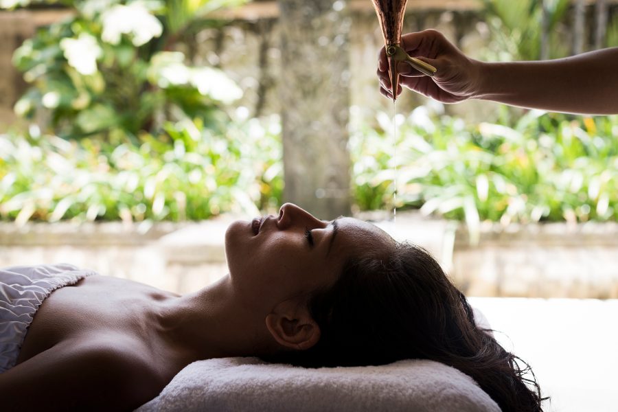 A woman lies on a massage table with her eyes closed as a massage therapist drips liquid onto her forehead at Revīvō Wellness Resort, Bali.