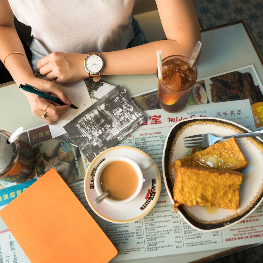 Person writing a note on a Hong Kong cafe table covered in Hong Kong-style milk tea, French toast and lemon tea. 