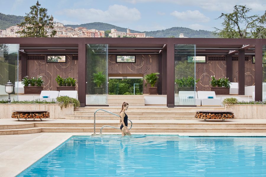 Woman in a black swimsuit descending the steps into a swimming pool in front of a set of luxury cabanas.