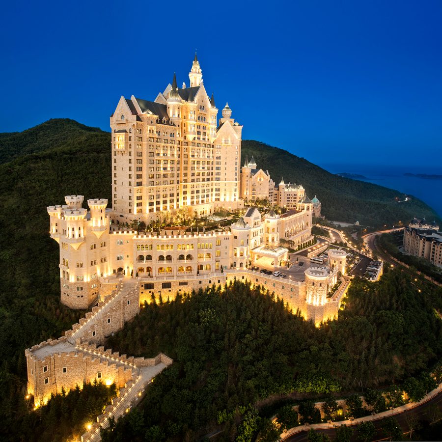 A drone shot of The Castle Hotel in Dalian, China, perched on top of a hill, lit up in warm lights after sunset