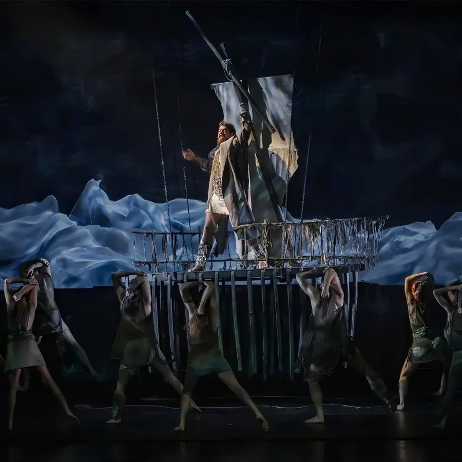 A man stands on a ship and sings to the crowd as performers create waves with cloth around him as part of an operatic version of The Divine Comedy.