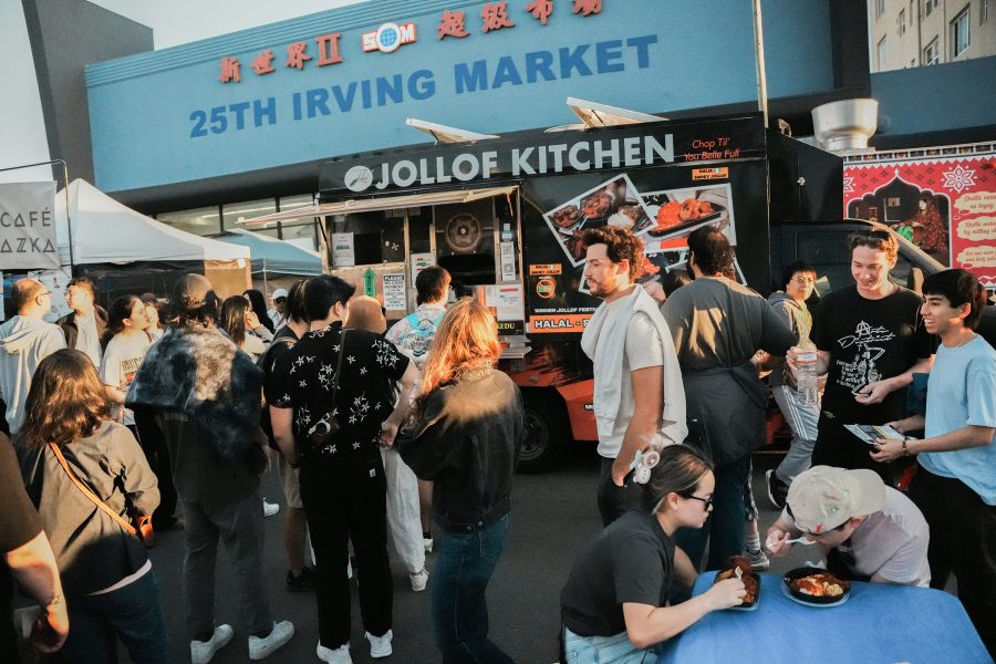 A long queue forms in front of a jollof kitchen food truck while people eat at a table on the side at San Francisco