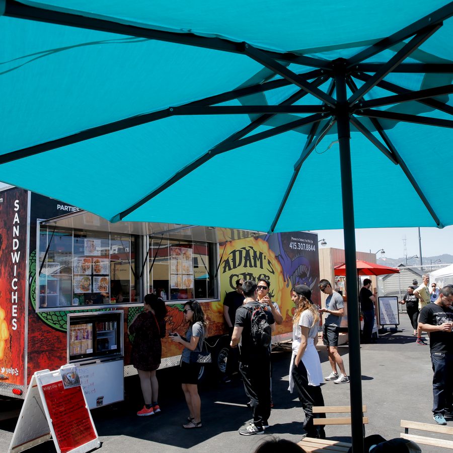 People lining up at a food truck under the sun with a blue umbrella in view