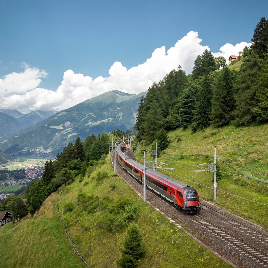 A sleek red-and-black train on the Brenner Railway hugs the mountain as it chugs onward.