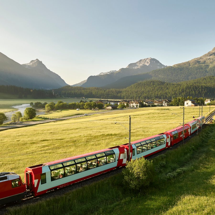 The Glacier Express is seen snaking through a pasture and heads for a mountain range in the distance. 