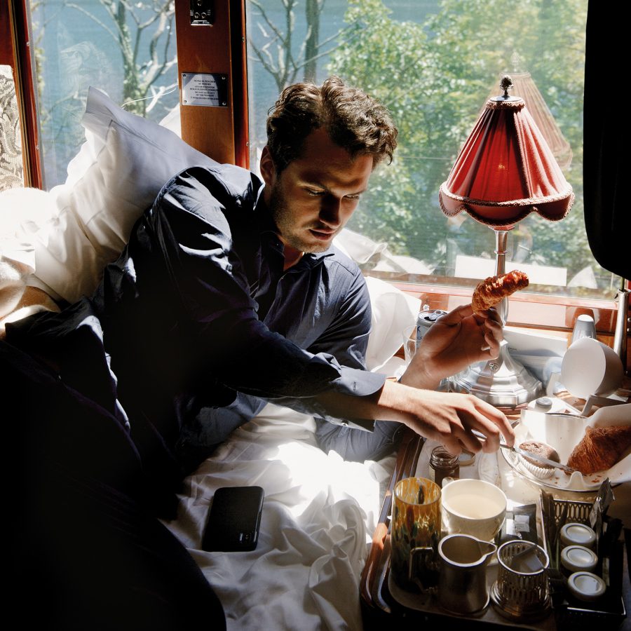 A male guest aboard the Venice Simplon-Orient Express lazing on pillows leans over the bedside table to eat breakfast as the scenery of the landscape passes by outside the window. 