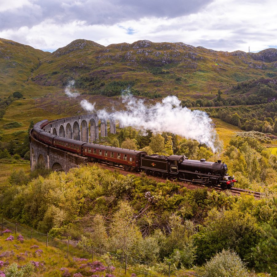A train on the West Highland Line expels smoke as it turns over a forested and mountainous landscape.