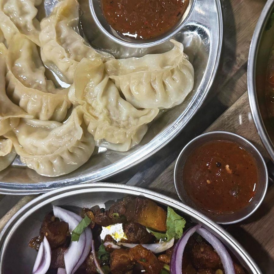 A large metal platter of momo dumplings with a bowl of red dipping sauce,  placed on wooden table surrounded by other dishes