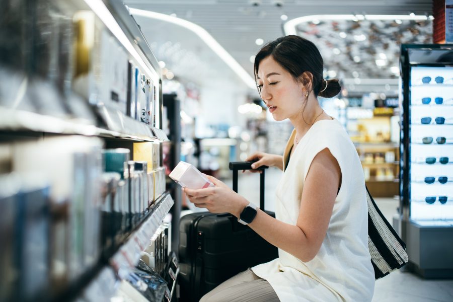 A woman with a suitcase kneels in front of a shelf at airport duty free to look at a perfume package