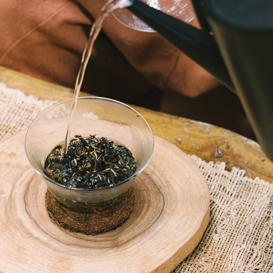 Person pouring hot water over tea leaves in clear glassware.