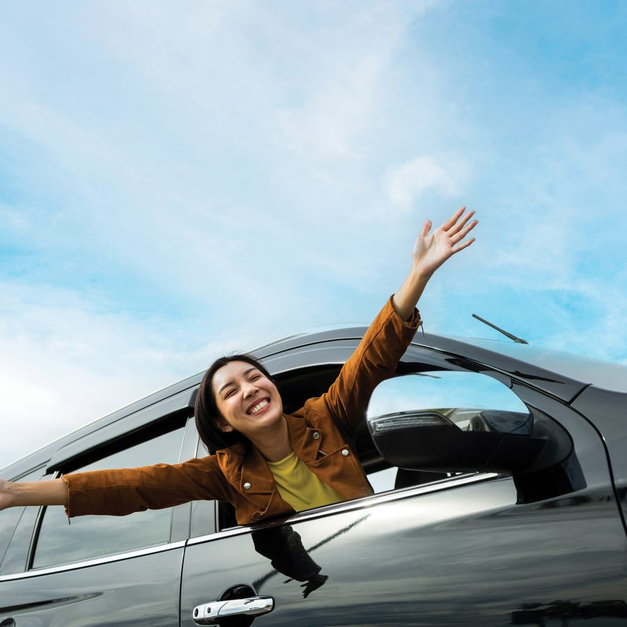 A smiling woman leans out of a car window with her arms outstretched.