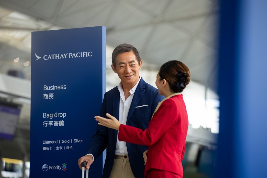 A smiling Asian man in a blue suit is shown to the Cathay Pacific Business check-in counter by a Cathay Pacific ground staff at Hong Kong International Airport. 