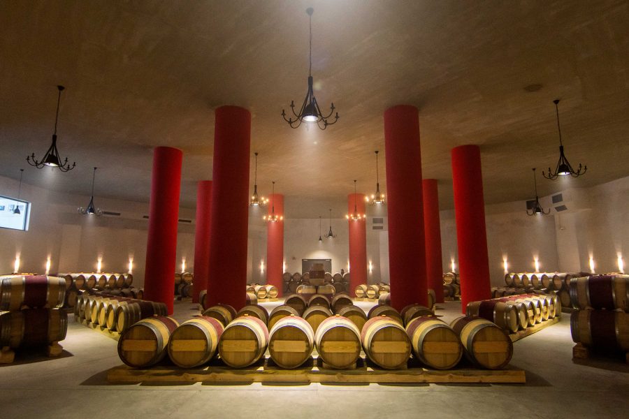 Wine barrels sit in a cellar ringed by crimson pillars and dangling chandeliers. 