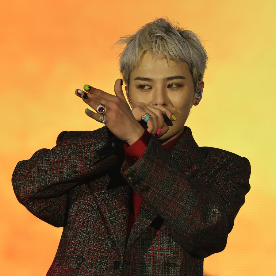 G-Dragon speaks into a microphone onstage against an orange backdrop. 