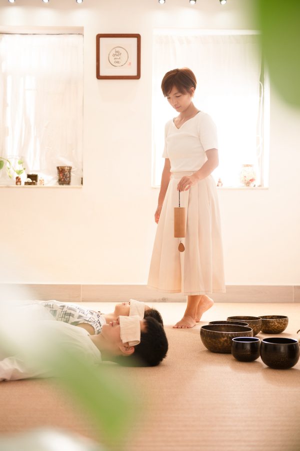A woman walking around a white room with singing bowls on the ground as people meditate. 
