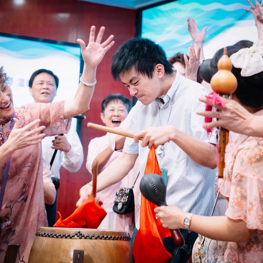 A young man plays the drums while a groups of older people stand around him cheering, in a scene from Big World.