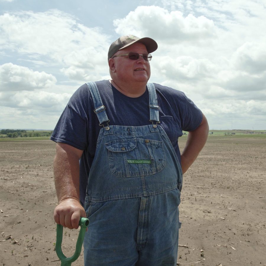 In a scene from Common Ground, a man dressed in overalls rests against a shovel in a large arid field.
