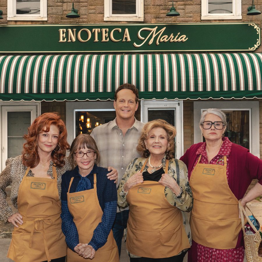 In a scene from Nonnas, a man and four older women wearing aprons stand smiling in front of an Italian restaurant.