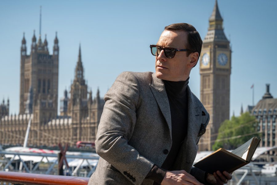 In a scene from Black Bag, a suave man in sunglasses stands on a bridge near the Houses of Parliament in London.