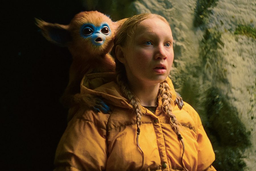 In a scene from The Legend of Ochi, a girl in a yellow jacket stands near a cave while a monkey-like creature sits on her shoulder.