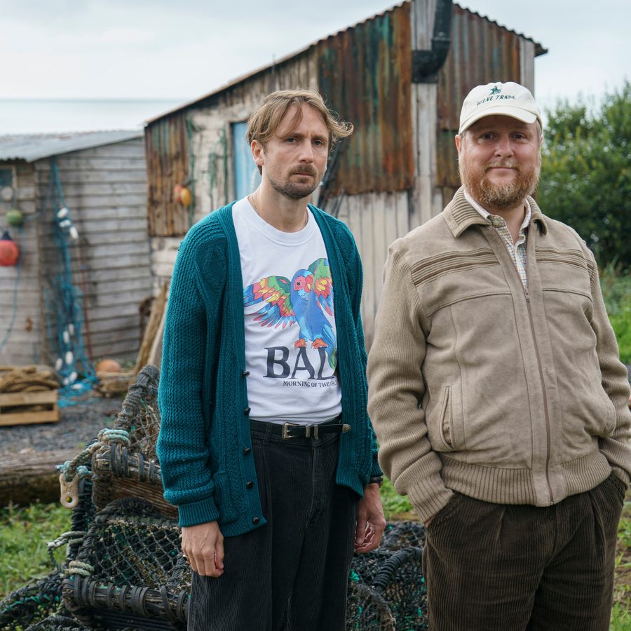 In a scene from The Ballad of Wallis Island, two men stand in front of an iron shed in a damp field.