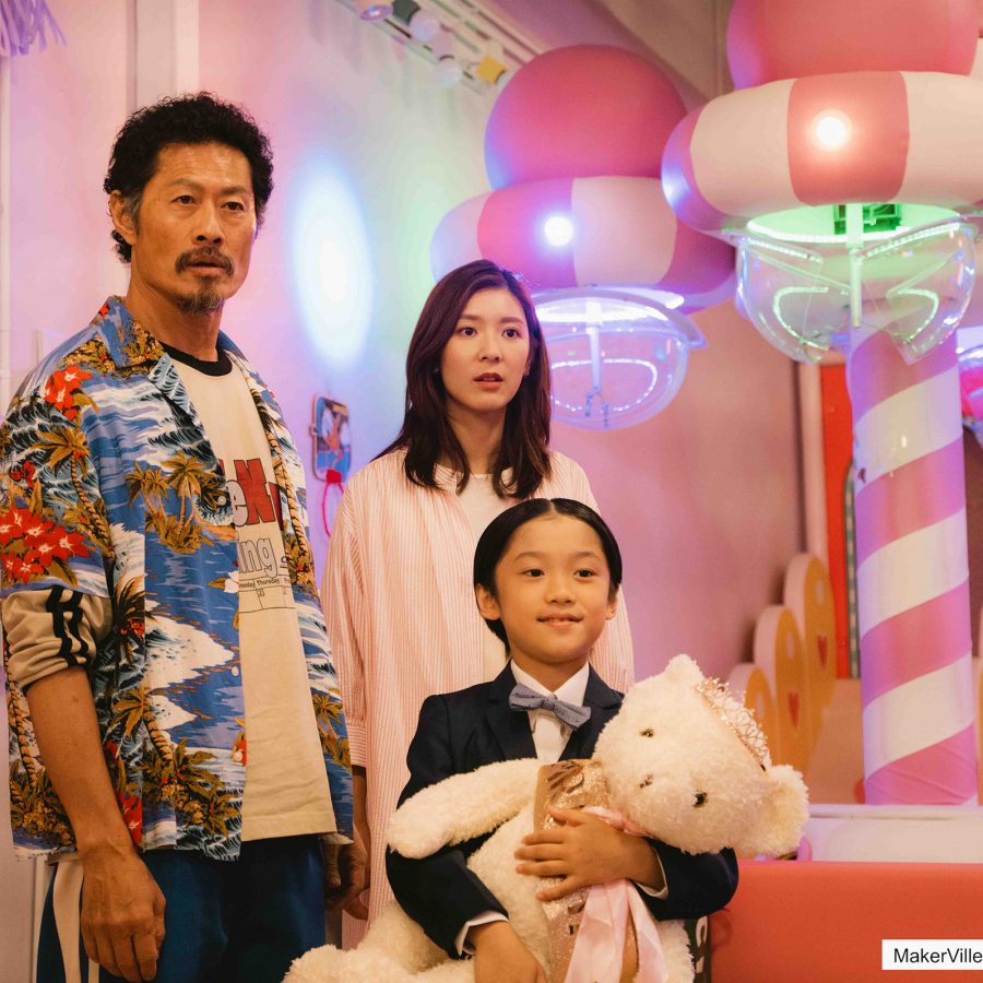 In a scene from Uncle Mum, an unkempt man and neat woman stand with a young girl holding a teddy bear in a pink room.